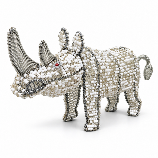 A rhinoceros figurine crafted from white and silver seed beads, featuring textured silver wire for the horns and legs, and a small red gemstone for the eye, set against a plain white background.