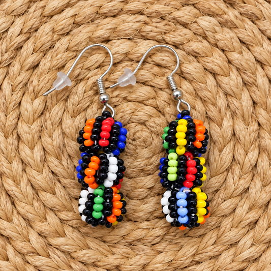 A pair of handmade beaded drop earrings with colorful seed beads in black, white, blue, red, yellow, green, and orange, hanging from silver fishhook wires on a circular woven straw mat.