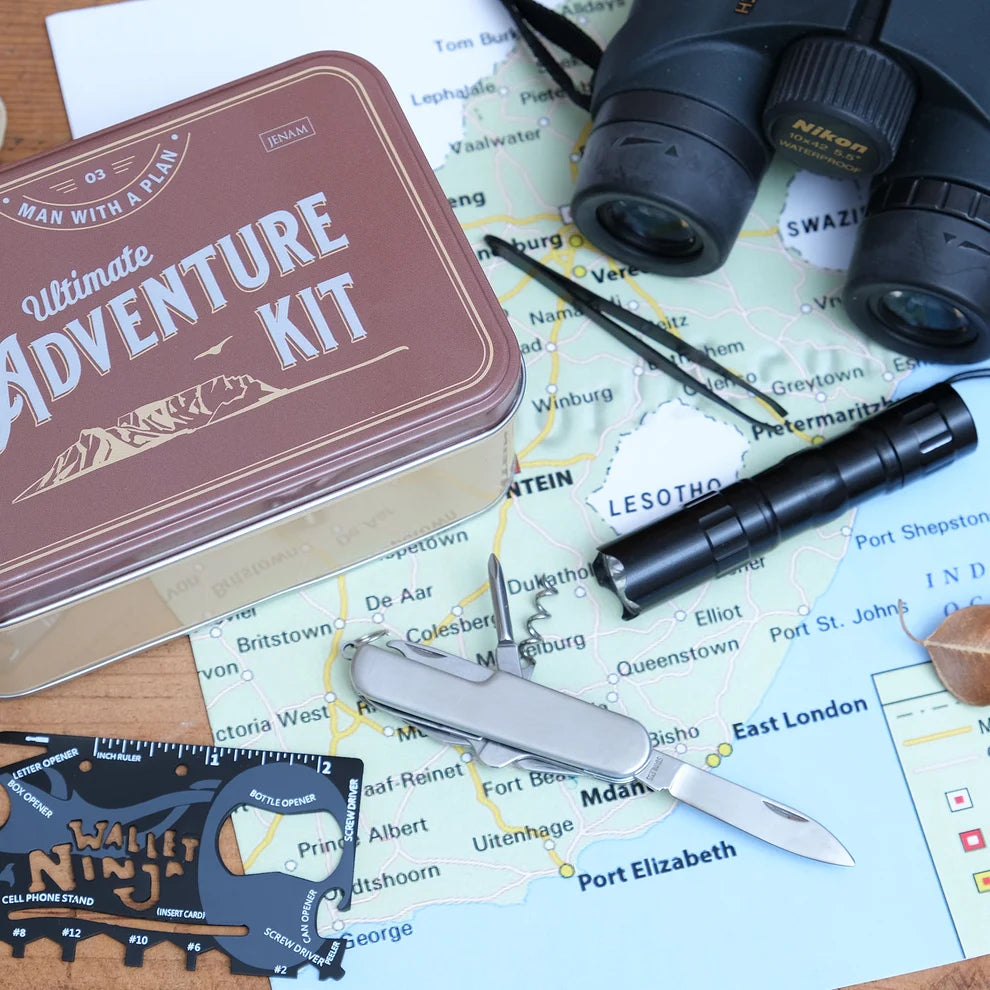 A family survival kit set out on a map, featuring a brown metal tin labeled 'Ultimate Adventure Kit', a black Nikon binoculars accessory, a black flashlight, a silver folding pocket knife, and a black family ninjaga cell phone stand on a world map.