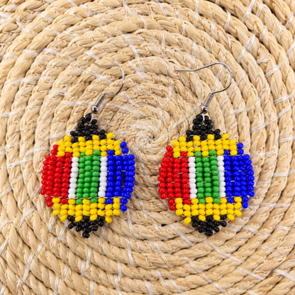 A pair of handmade beaded drop earrings featuring a multicolor striped design inspired by the South African flag, displayed on a woven straw mat.