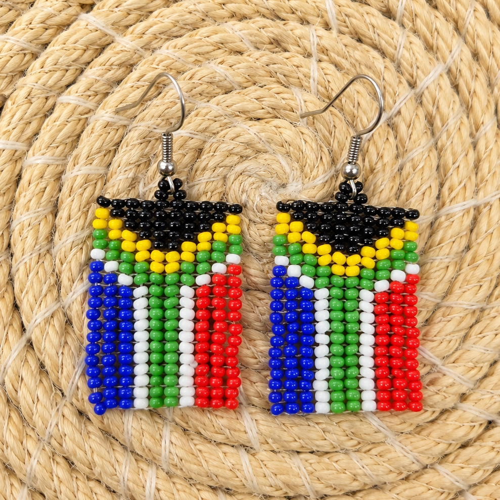 A pair of handmade beaded earrings featuring the South African flag design, displayed on a circular woven straw mat. The earrings have silver fishhook wires and are made of seed beads in black, yellow, green, white, and red.
